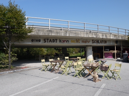 Tische und Stühle vor und unter einer Autobrücke bilden ein temporäres Wirtshaus, dazu der Schriftzug "eine Stadt kann nicht mehr schlafen" an der brücke